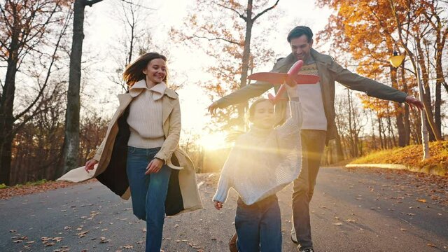 Young parents laughing, running by road with little daughter holding red toy plane. Autumn park. Happy family. Close up, slow motion. Tracking shot