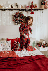 Little girl child in red pajamas jumping on the bed and enjoying the Christmas holidays. A child in an apartment with a Christmas decoration is having fun. Happy childhood and New Years celebration