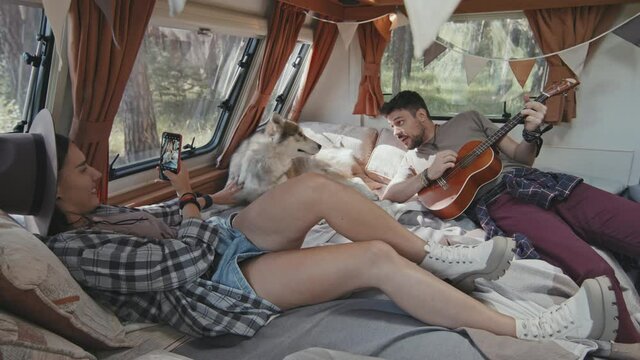 Tracking shot of happy young woman and man with ukulele relaxing on bed in cozy camper. Woman smiling and taking photos of adorable husky dog