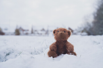 Lost Teddy bear with sad face lying on snow with blurry house,Lonely bear doll sitting alone on the...