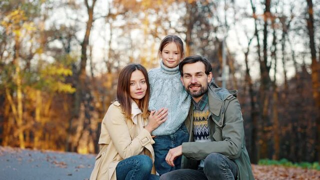 Young smiling mother and father sitting squatting in autumn park and hugging their little daughter. Happy family. Close up, slow motion