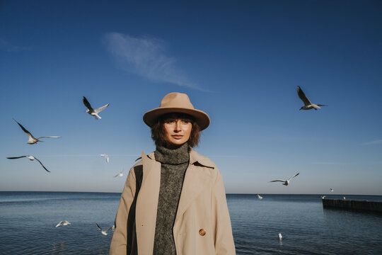 Woman in overcoat and hat standing in front of seagulls flying over sea