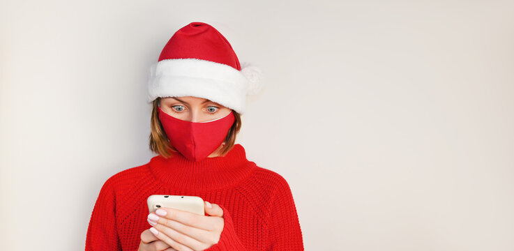 Christmas Woman in santa's hat, red sweater and wearing red medical nask stares surprised at phone's screen. Online shopping.Sale and discount concept.Mockup