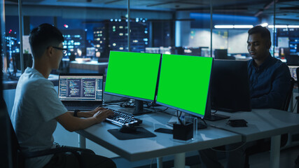 Night Office: Young Japanese Man in Working on Green Screen Chroma Key Desktop Computer. Team of Programmers Typing Code, Creating Modern Software, e-Commerce App Design, e-Business Programming