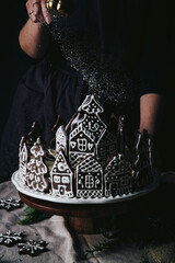 Christmas gingerbread village dusted with powdered sugar. Homemade two-tier cake decorated with hand painted gingerbread cookie houses and trees on wooden stand on dark background .