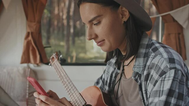 Handheld close up with tilt up of young woman in hat holding ukulele and sitting on bed in camper while typing on mobile phone