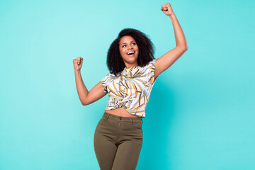 Photo of charming lucky dark skin woman dressed print blouse rising fists looking empty space smiling isolated teal color background