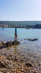 Fototapeta na wymiar A young man in a biking outfit, standing on a rock in a shallow sea water and admiring the view in front of him. Water has many shades of blue. Sea is very calm. Clear and sunny day.