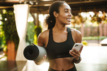 Black woman smiling and using cellphone while standing with yoga mat
