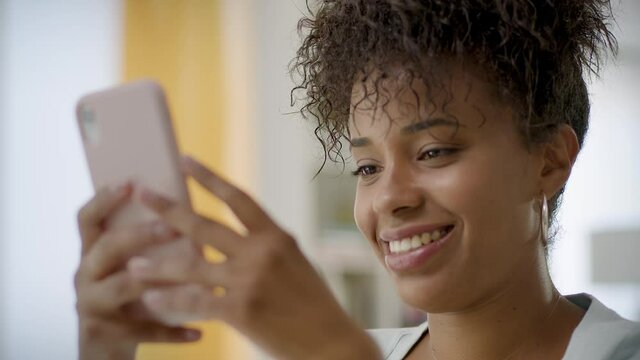 Lateral Dolly of Cheerful Young African American Woman Using Smartphone at Home or Office. Black Smiling Woman texting on Cellphone. Beautiful Girl Chatting on Mobile Phone.