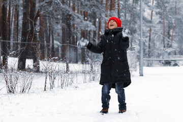 A boy in a red hat happily throws snow up in the winter forest