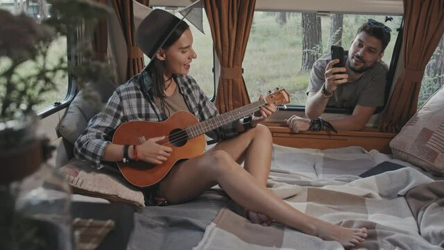 Medium shot with slowmo of cheerful man looking through open window of camper and filming happy young woman sitting on bed inside and playing ukulele, then giving her high-five