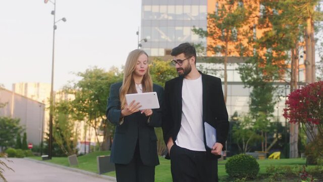Walking in front of the camera two charismatic students lady and guy discussing holding a electronic tablet and analysing something background modern college building