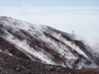 Silhouette of man hiking at coldera of Avachinsky stratovolcano, also known as Avacha Volcano. Backpacker tourist moves upon rocks behind steam from hot geysers. Kamchatka Peninsula, Russia.