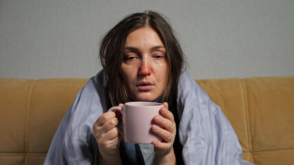 Young woman with long hair and runny nose falls ill and drinks hot tea from mug wrapped in warm blanket, sitting on comfortable sofa in living room.