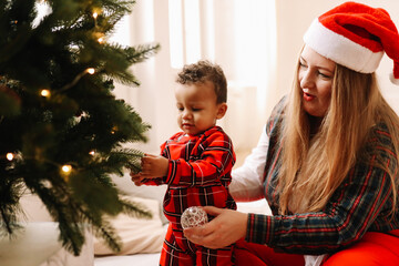 Multiracial mom and small child son in red plaid pajamas have fun decorating the Christmas tree during the winter holidays in a cozy bright room at home in December
