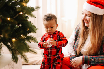 Multiracial mom and small child son in red plaid pajamas have fun decorating the Christmas tree during the winter holidays in a cozy bright room at home in December