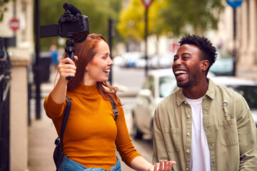 Young Couple Travelling Through City Together Vlogging To Video Camera On Handheld Tripod