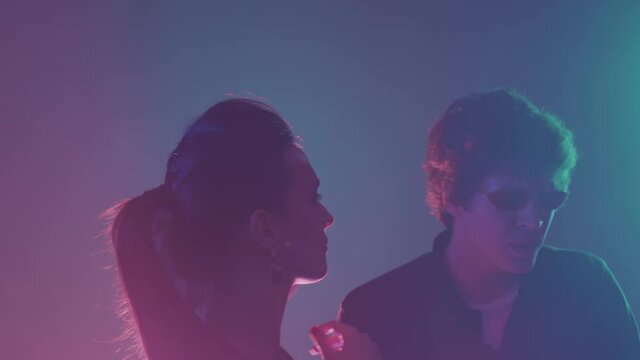 Medium close up with slowmo of handsome guy in trendy sunglasses dancing with two beautiful girls at party in night club on neon lights