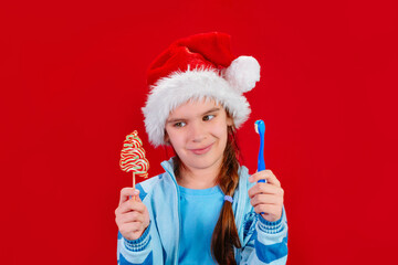 Happy Christmas. Girl kid in a Santa's hat with a candy and a blue toothbrush in his hands on red background. To choose. Place for text. Dental hygiene, children's health concept.