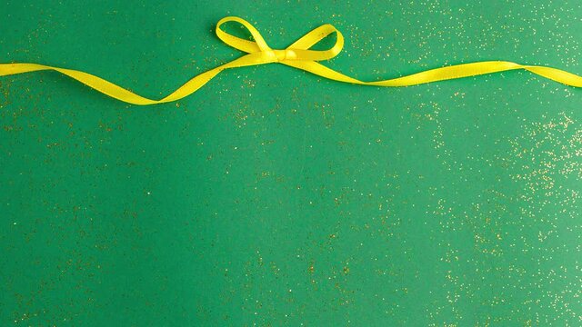A yellow satin ribbon bow tied on jade green background with gold colored glitter. Stop motion animation Christmas Holidays and present concept flat lay with copy space