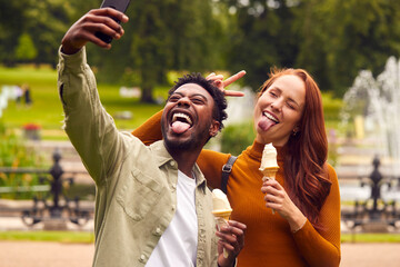 Obraz premium Young Couple Travelling Through City Together Eating Ice Creams In Park And Taking Selfie