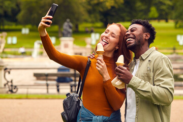 Young Couple Travelling Through City Together Eating Ice Creams In Park And Taking Selfie