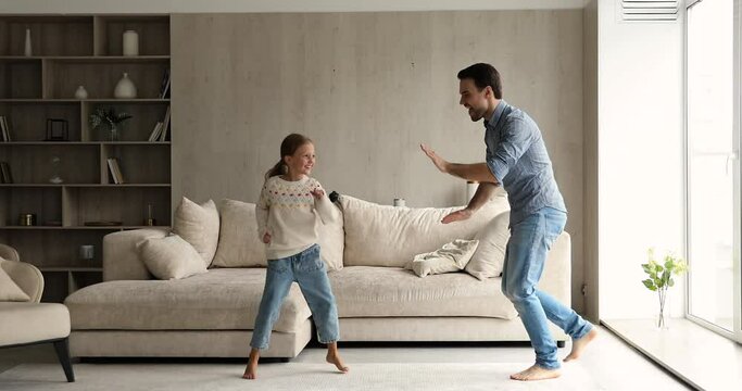 Dances in living room. Joyful young father and small daughter have fun jump play move by active music celebrate relocation to new modern apartment. Adult elder brother dancing with younger sister kid