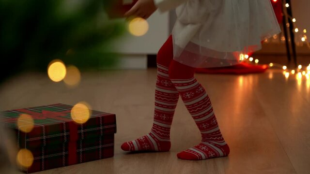 Little childrens feet in red pantyhose with New Years ornament go to Christmas tree with gifts at dark. Child in socks puts gift under Christmas tree