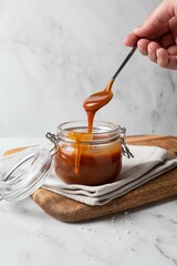 Homemade salted caramel sauce in glass jar on wooden board, grey background. Caramel sauce drips from spoon in hand.