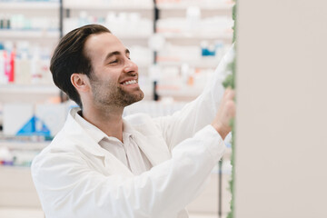 Smiling male young caucasian pharmacist druggist stretching to the shelves with medicines drugs, antibiotics, painkillers, vitamins at pharmacy drugstore