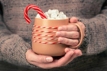 Close up of female hands in warm wool sweater holds brown cozy mug with hot cocoa, chocolate or coffee with marshmallow and candy cane. Christmas time concept.