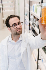 Male young caucasian pharmacist druggist in medical white coat stretching to pills, vitamins, painkillers, medicines at pharmacy shelves in drugstore