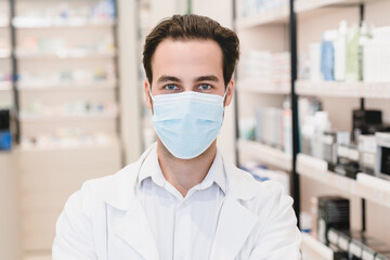 Fototapeta na wymiar Pandemic, lockdown. Caucasian male young pharmacist druggist in white medical coat and protective face mask against Covid19 looking at camera in pharmacy drugstore