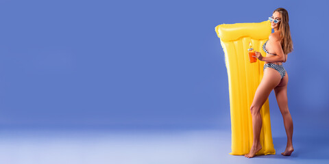 Back view of young woman posing in bikini with yellow inflatable mattress while holding cocktail. 