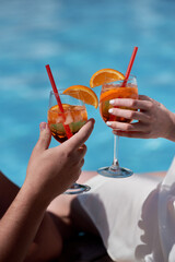Couple hands toasting with aperol spritz cocktails on summer party by the pool, copy space. Event celebration concept. Traditioanal italian aperitif.