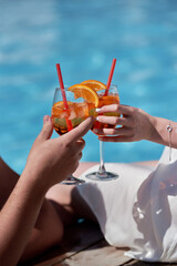 Couple hands toasting with aperol spritz cocktails on summer party by the pool, copy space. Event celebration concept. Traditioanal italian aperitif.