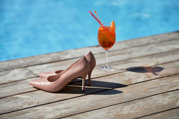 Aperol spritz cocktail and beige woman shoes by the pool, copy space. Event celebration concept....