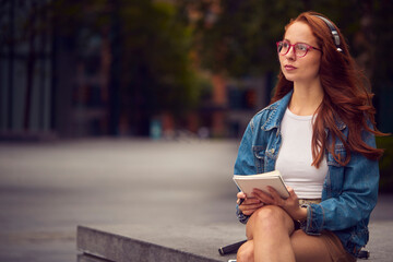 Female Vlogger Or Social Influencer Wearing Headphones Travelling Through City Making Notes In Book