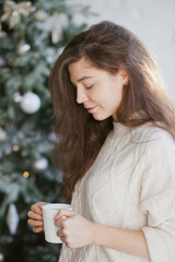 Beautiful young woman drinking coffee or tea near christmas tree in the living room at home close up