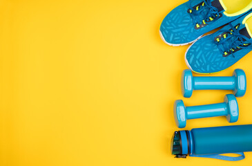 Blue dumbbells, trainers and a water bottle on yellow background, top view with copy space