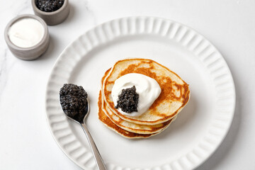 Black Caviar on pancakes with sour cream Holiday morning Appetizer on white plate and marble background with spoon of caviar - obrazy, fototapety, plakaty