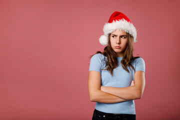 Angry unhappy caucasian girl teenager standing in santa claus red hat looking aside over pink background. Studio portrait. Anti Christmas and New year Holiday concept with copy space.