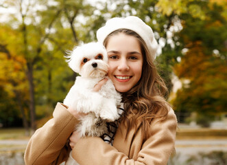 Portrait of young happy Caucasian teenager girl in french white hat and warm coat holding and hugging her little cute white dog maltese mini in autumn park