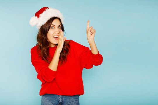 Young happy woman with curly hair in Santa Claus hats standing over blue background smiling with happy face looking and pointing to the side with thumb up. Place for your text