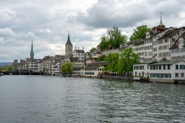 Fototapeta premium The Limmat River flows next to the cityscape of Zurich, Switzerland on a cloudy spring afternoon - trees and buildings line the river