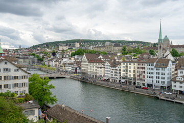 Fototapeta na wymiar The Limmat River flows next to the cityscape of Zurich, Switzerland on a cloudy spring afternoon - trees and buildings line the river