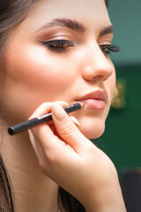 Makeup professional artist or cosmetologist is painting contour lips of a young woman with a pencil close up