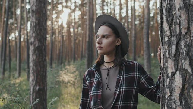 Tracking shot of pensive young woman in hat standing in tranquil forest and looking away from camera