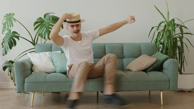 Funny modern fashion hipster with mustache, straw hat, round pink sunglasses chilling, fooling around, lying on sofa while dancing in modern loft apartment. Millennial killing time lifestyle
